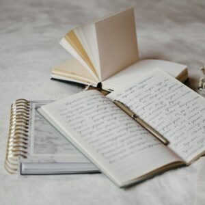 Close-up of open notebooks and a pen on a soft bed surface with delicate dried flowers.